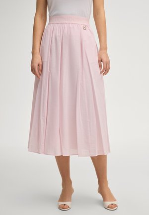 Lichtroze geplooide midi-rok met een aansluitende tailleband, gecombineerd met witte open teen hakken, gedragen door een staand persoon tegen een effen achtergrond.