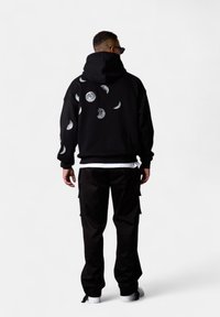 Haut à capuche noir avec des graphiques des phases de la lune au dos ; coupe décontractée, poignets et ourlet côtelés. Porté avec un pantalon cargo noir et des baskets blanches.