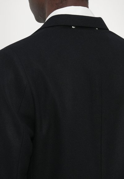 Sacou negru, croit pe măsură, cu un material texturat, rever ascuțit și un detaliu decorativ subtil pe spate, cu un mic accent metalic.