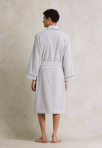 Άτομο όρθιο, ξυπόλυτο, φοράει ανοιχτογκρίζο μπουρνούζι μήκους μέχρι το γόνατο με ζώνη και μαύρο περίγραμμα στα μανίκια, με την πλάτη γυρισμένη προς ουδέτερο φόντο.