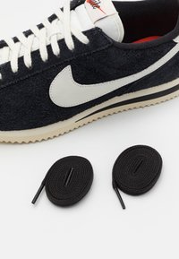 Musta mokkanahkainen Nike-lenkkiurheilukengät, valkoisella swoosh-logolla ja nauhoilla, esiteltynä kierrettyjen mustien nauhojen kanssa valkoista taustaa vasten.