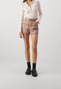 Haut en tricot à manches longues couleur crème avec patte de boutonnage, associé à un short taille haute à carreaux marron et violet, doté d'une ceinture et de poches.