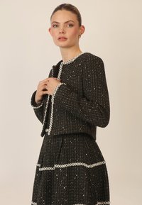 Chaqueta de tweed negra con acentos de perlas, con cuello redondo y mangas largas, combinada con una falda a juego con diseño en capas.
