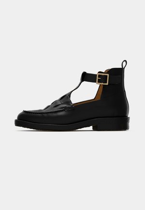 Scarpe in pelle nera con un design a T, punta tondeggiante, dettagli intrecciati e tacco basso. Presentano una fibbia metallica per la chiusura.