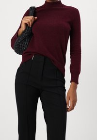 Pull en maille bourgogne avec un col montant, associé à un pantalon noir ajusté et un sac à main noir texturé. Textures lisses et douces dans l'ensemble.