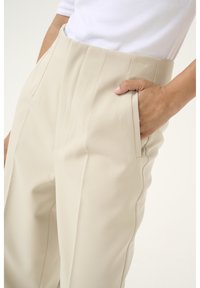 Pantalons beige ajustés en tissu texturé avec des plis avant et des poches latérales, dotés d'une taille haute et d'une finition soignée.