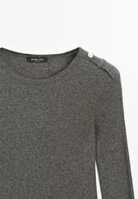 Maglione grigio con scollo rotondo, texture a coste e bottoni decorativi argentati sulla spalla sinistra. Materiale morbido con un design aderente.
