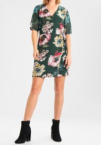 Vestido envolvente floral en verde oscuro con mangas cortas. Presenta patrones florales en rosa, blanco y azul. Combinado con botas de tobillo negras.