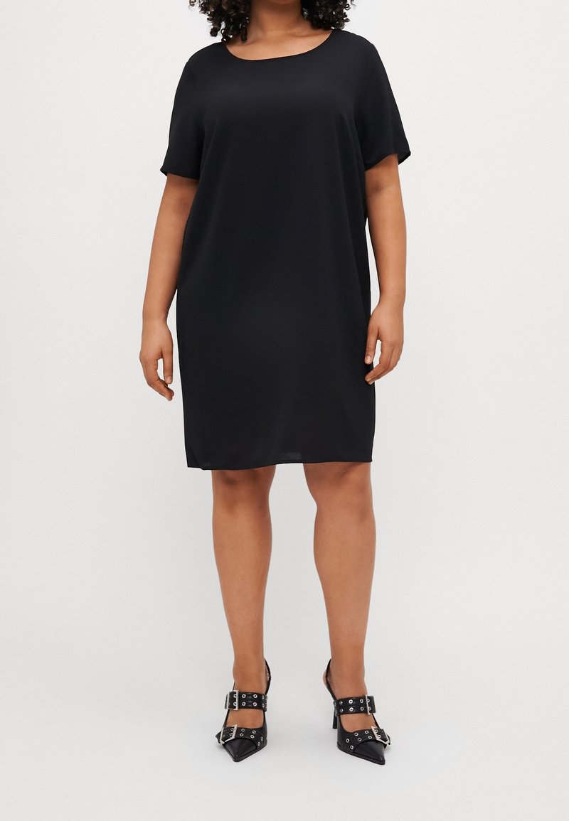 Vestido negro de manga corta con un ajuste relajado y cuello redondo, confeccionado en una tela suave. Se lleva con zapatos negros de punta con detalles de hebilla.