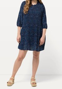 Kvinna klädd i en marino-blå blommig plisserad klänning med 3/4-långa ärmar och beige platåsandaletter, stående mot en enkel vit bakgrund.