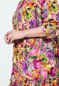 Květinové šaty s živými barvami růžové, fialové a oranžové; mají zvýrazněný pas, knoflíkové detaily a krátké rukávy s ohrnutými manžetami.