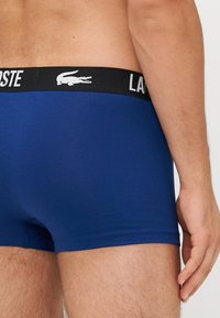 Bóxer azul marino con una cinturilla elástica negra que presenta el texto y el logo en blanco "LACOSTE", fabricado con una tela suave y elástica.