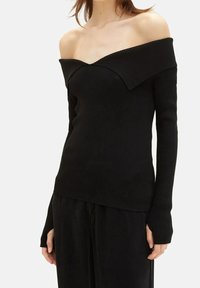 Femme portant un haut noir côtelé à manches longues échancré aux épaules et un pantalon noir ample, debout devant un fond clair uni.