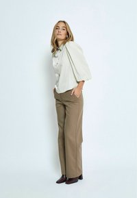 Light beige puff-sleeve shirt with buttons, paired with high-waisted, tailored brown pants and dark block-heeled boots.