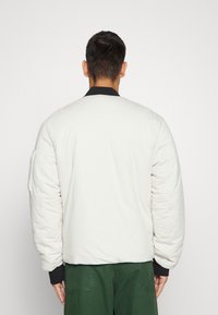 White padded bomber jacket with a black collar, featuring ribbed cuffs and a side pocket, displayed from the back.