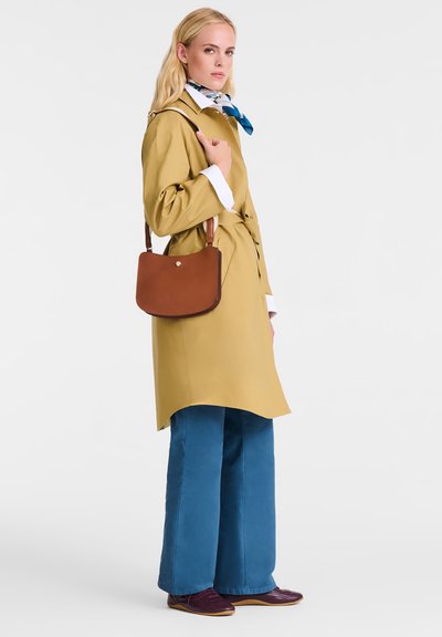 Frau trägt einen senfgelben Trenchcoat, weite blaue Hose, bordeauxfarbene Schuhe, ein blau-weißes Halstuch und eine braune Schultertasche.