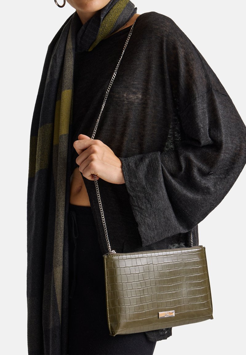 Borsa a mano verde oliva con stampa coccodrillo, hardware argentato e tracolla a catena, indossata con un maglione oversized nero e una sciarpa grigia a righe.