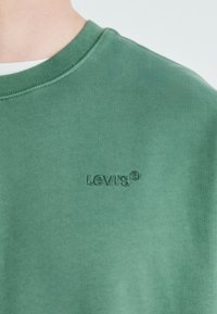Sudadera verde de algodón con cuello redondo de canalé; cuenta con un logo de Levi's en relieve en el lado izquierdo. Textura suave, diseño casual.