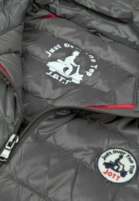 Chaqueta negra acolchada con cremallera plateada, logo interior en blanco y rojo que muestra a una persona montando un patinete y el texto "Just Over The Top JOTT."