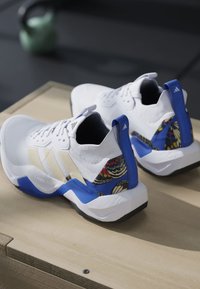Scarpe sportive bianche con accenti blu, strisce beige e motivi colorati di farfalle sul tallone, posizionate su una superficie in legno.