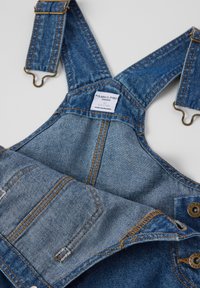 Farkkuhaalarit sinisestä kankaasta, säädettävillä olkahihnoilla, metalliklipsuilla ja kultaompeleilla. Sisältää etutaskun ja merkkilapun.