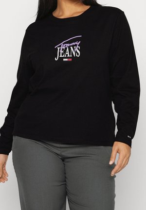 Sort langærmet skjorte med "Tommy Jeans" logo i hvidt og lilla over et lille rødt, hvidt og blåt flag, båret med grå bukser.