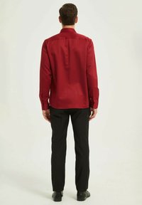 Chemise rouge à manches longues avec col boutonné, texture lisse, et pantalon noir. La chemise a un ourlet droit et des poignets boutonnés.