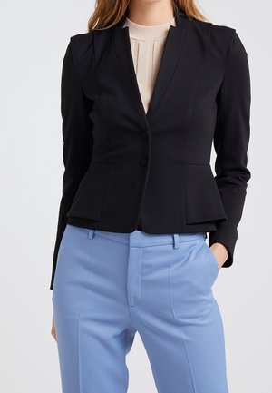 Vrouw draagt een nauwsluitend zwart blazer over een beige top, gecombineerd met hoog getailleerde lichtblauwe broek, één hand in de zak.