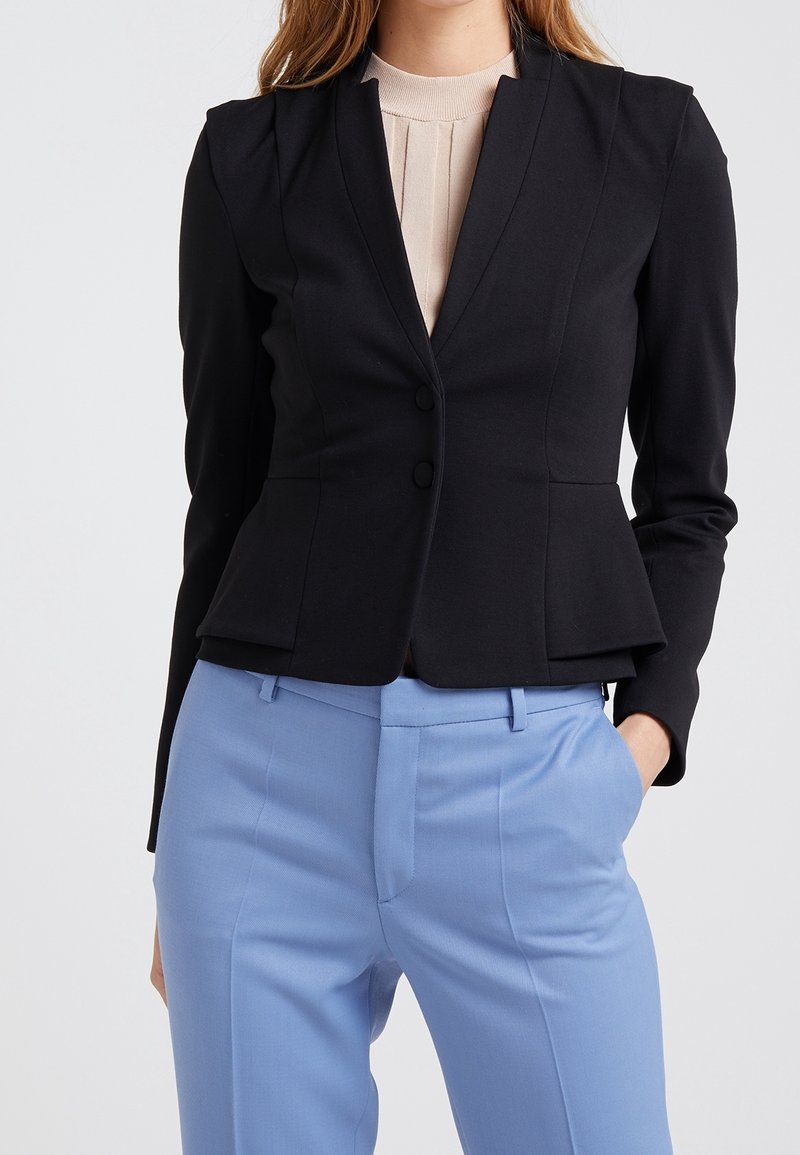 Femme portant un blazer noir cintré sur un top beige, associé à un pantalon taille haute bleu clair, une main dans la poche.