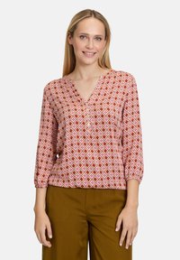 Blouse à motifs avec une base rose, présentant des formes géométriques en rouge et jaune, décolleté en V et manches trois-quarts. Associée à un pantalon marron.