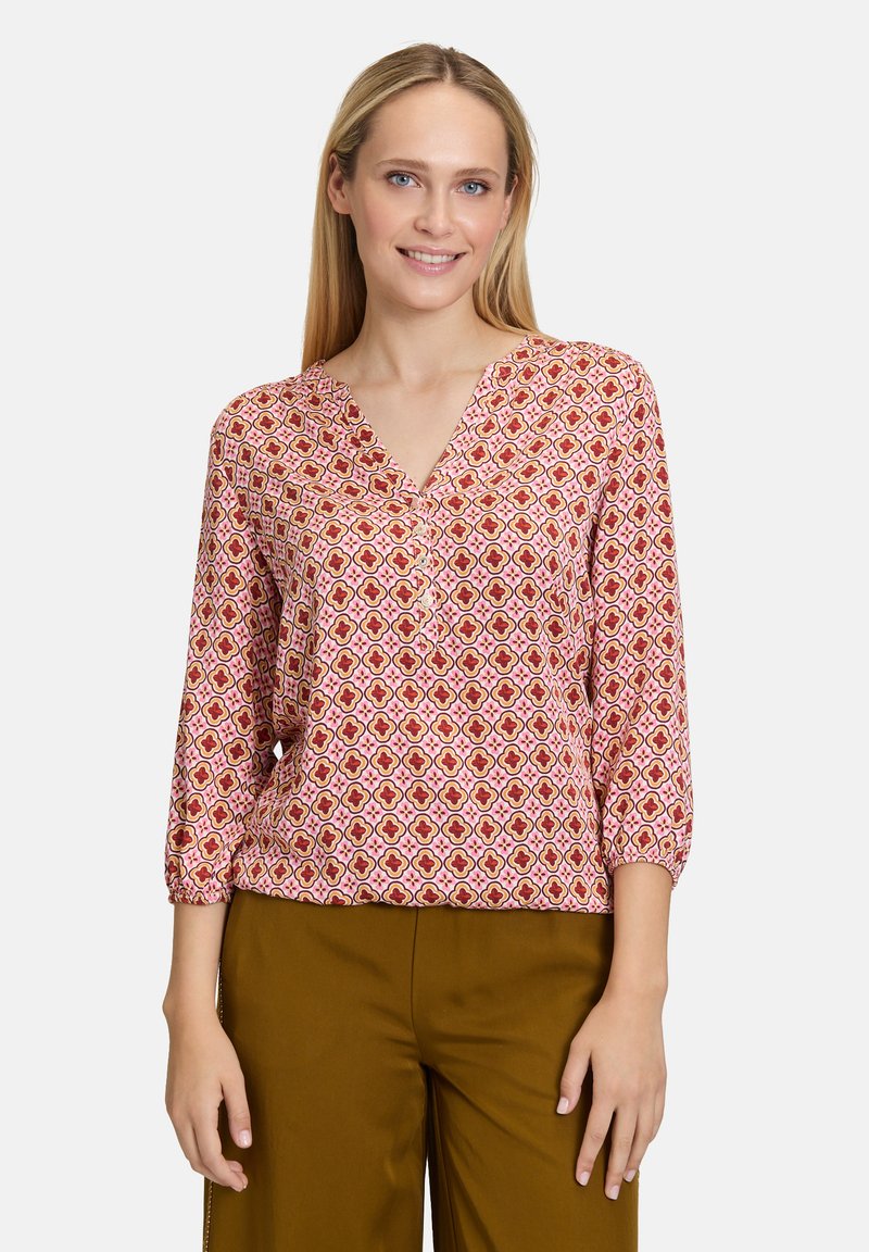 Blouse à motifs avec une base rose, présentant des formes géométriques en rouge et jaune, décolleté en V et manches trois-quarts. Associée à un pantalon marron.