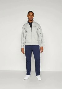 Casaco cinza com fecho, com detalhes em preto, combinado com calças azul-marinho e ténis brancos. Tecido suave, design ajustado e punhos canelados.