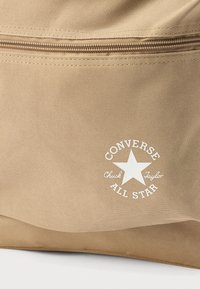 Τσάντα από καφέ ύφασμα με υφή στην επιφάνεια, που διαθέτει ένα λευκό κυκλικό λογότυπο με ένα αστέρι και το κείμενο "CONVERSE CHUCK TAYLOR ALL STAR." Κλείσιμο με φερμουάρ.