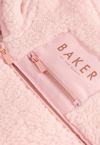 Blødt pink tekstureret stof med to rosaguld lynlåse og en "BAKER by Ted Baker" label på et tøjstykke.