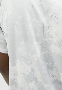 Vaaleanharmaa urheilu T-paita, jossa on teksturoitu kuvio ja hienovaraisia tummempia läiskiä. Sisältää sivupaneelin ja lyhyet hihat.