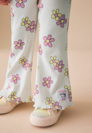 Vaikas dėvi baltas kelnes su rožinėmis ir geltonomis gėlių spaudomis bei rožiniais batais, stovintis ant smėlio spalvos fono.