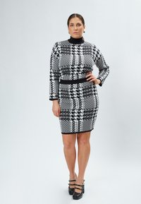 Strikket kjole i sort og hvid geometrisk mønster; tætsiddende facon med lange ærmer, høj hals og taljeaccent. Matchet med sorte højhælede sko.