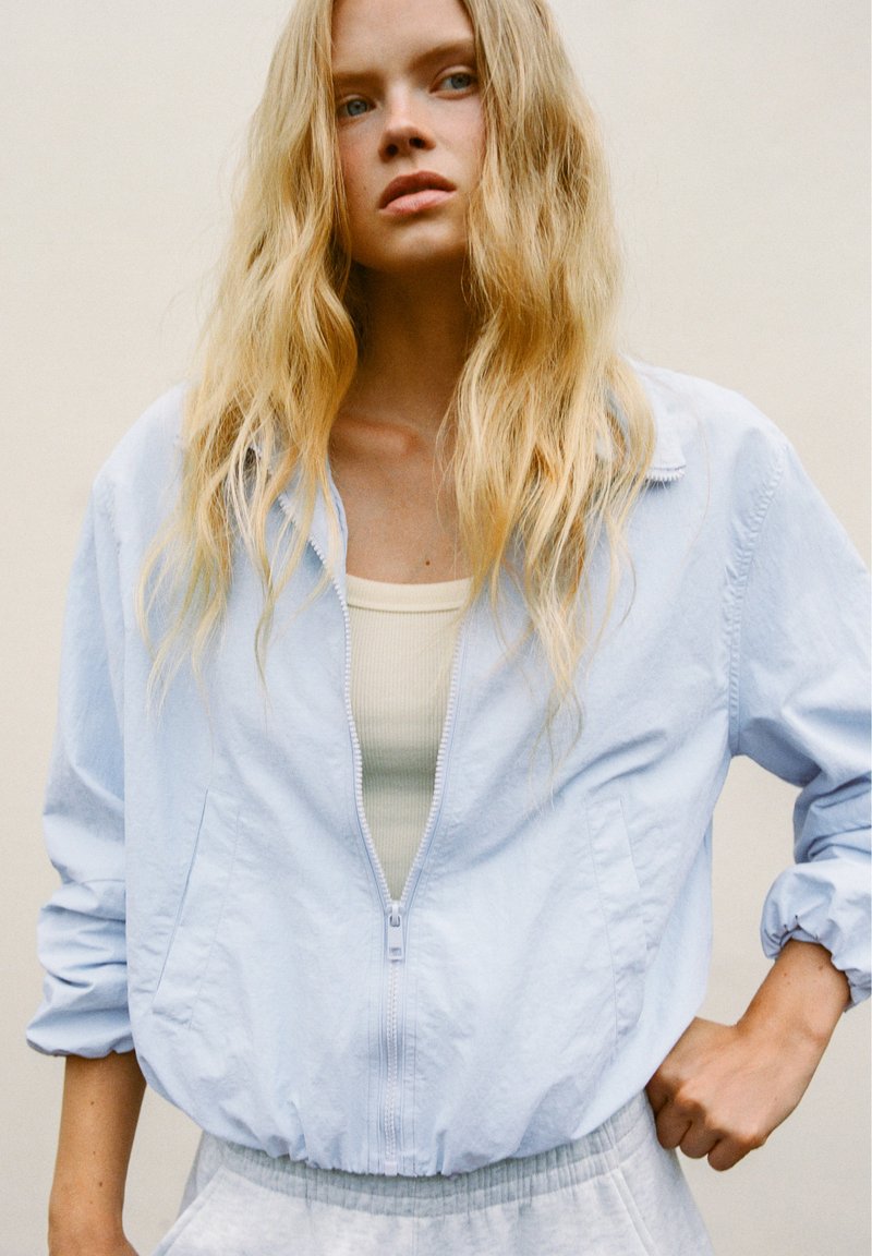 Femme blonde aux cheveux ondulés portant une veste zippée bleu clair sur un top beige, debout, une main sur la hanches, devant un fond uni.
