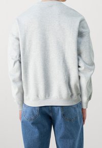 Osoba ubrana w jasnoszary sweatshirt i niebieskie dżinsy, pokazana od tyłu na jednolitym jasnym tle.