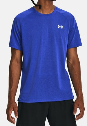 Mann trägt ein königsblaues, kurzärmeliges Sportshirt mit weißem Under Armour-Logo auf der Brust und schwarze Shorts, steht vor weißem Hintergrund.