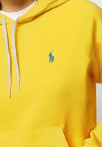 Gul huvtröja i tyg med en slät yta, med en blå logotyp av en häst med en ryttare. Vita snören och en framficka.