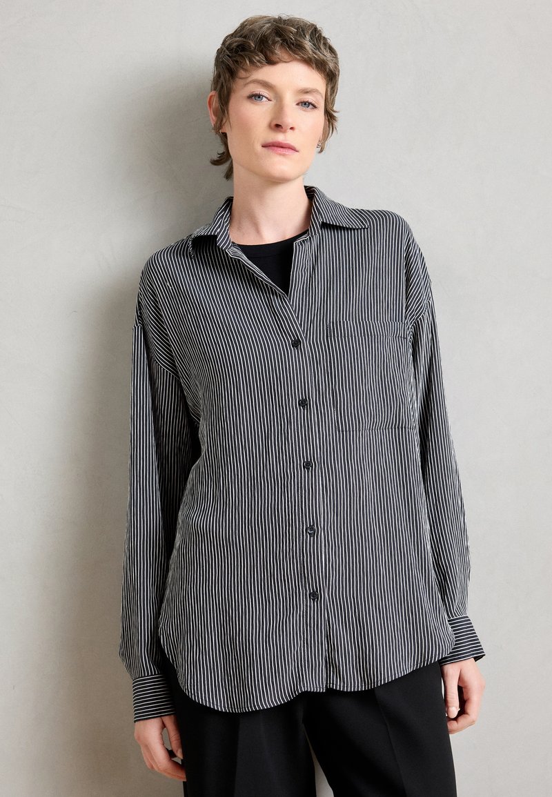 Camicia a righe bianche e nere con colletto e maniche lunghe. Presenta una tasca sul petto e un orlo arrotondato.