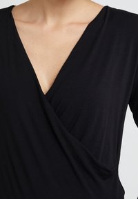 Schwarzes Wickel-Oberteil aus weichem Material, mit tiefem V-Ausschnitt und drapiertem Design an der Vorderseite. Glattes und tailliertes Design.