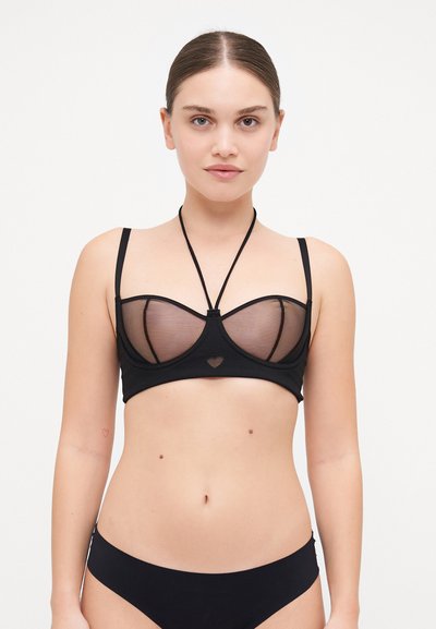 Bralette noire avec des panneaux en mesh transparent, découpe en forme de cœur au centre, bretelles réglables et design ajusté. Esthétique minimaliste.