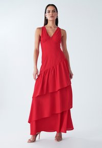 Red maxi dress featuring a V-neckline, layered ruffled design, and a flowing silhouette. Made from lightweight fabric, worn with strappy heels.