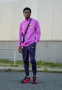 Nike sportoutfit met een paarse jas met logo en navy broek. Zwart-rode sneakers, vergezeld van een kleine zwarte schoudertas.