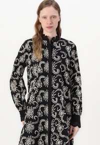 Vestido preto bordado com intrincados padrões florais brancos, mangas longas com punhos de renda e um design com botões.