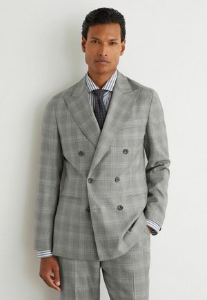 Man wearing a light gray double-breasted suit with a striped shirt and dark patterned tie, standing against a white wall.