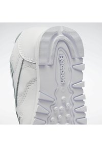 Fehér sneaker, textúrált bőrből készült felsőrésszel és szürke díszítéssel. Gumitalppal rendelkezik, amely bordázott mintázattal bír, és az alján a "Reebok" logó található.