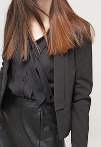 Blazer nero con design aderente, abbinato a una blusa nera con un nodo al collo e una gonna di pelle, il tutto in un look elegante.